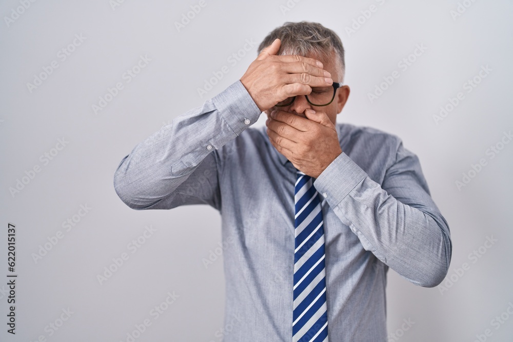 Hispanic business man with grey hair wearing glasses covering eyes and mouth with hands, surprised and shocked. hiding emotion Hispanic business man with grey hair wearing glasses covering eyes and mouth with hands, surprised and shocked. hiding emotion