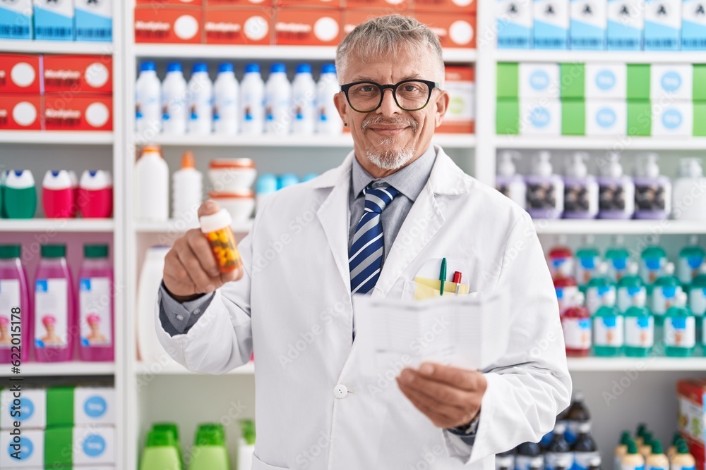 Middle age grey-haired man pharmacist holding pills bottle reading prescription at laboratory