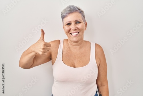 Middle age caucasian woman standing over white background doing happy thumbs up gesture with hand. approving expression looking at the camera showing success.