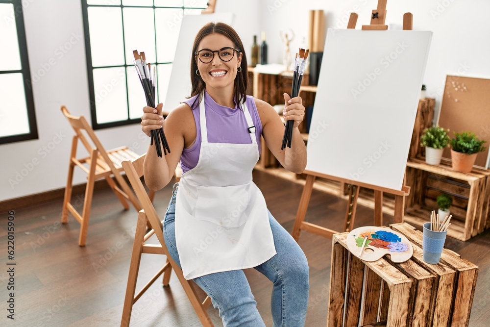 Young beautiful hispanic woman artist smiling confident holding paintbrushes at art studio