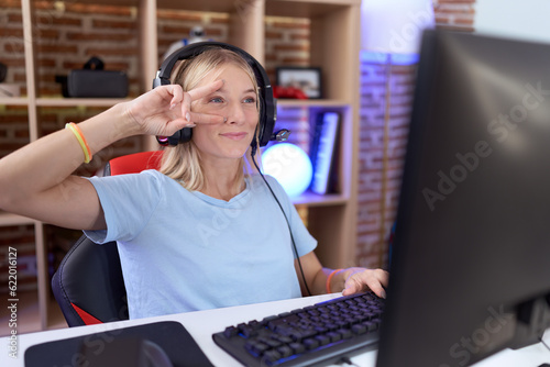 Young caucasian woman playing video games wearing headphones doing peace symbol with fingers over face, smiling cheerful showing victory