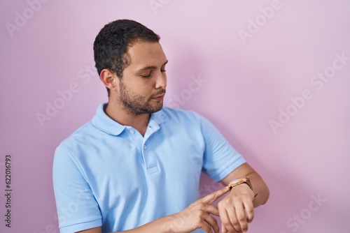 Hispanic man standing over pink background checking the time on wrist watch, relaxed and confident