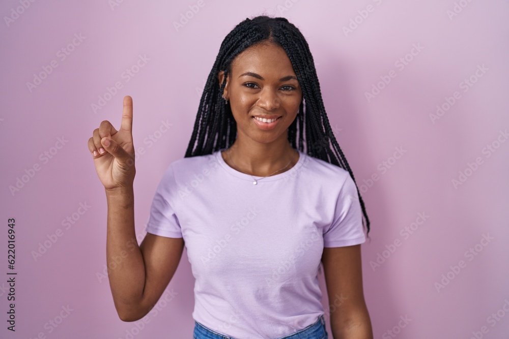 African american woman with braids standing over pink background showing and pointing up with finger number one while smiling confident and happy. African american woman with braids standing over pink background showing and pointing up with finger number one while smiling confident and happy.
