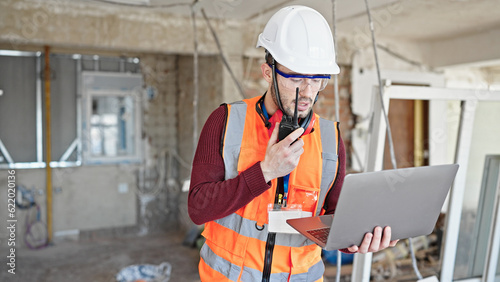 Young hispanic man builder talking on walkie talkie using laptop at construction site