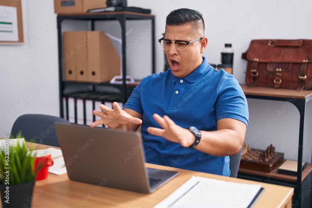 Young hispanic man working at the office with laptop afraid and terrified with fear expression stop gesture with hands, shouting in shock. panic concept. Young hispanic man working at the office with laptop afraid and terrified with fear expression stop gesture with hands, shouting in shock. panic concept.