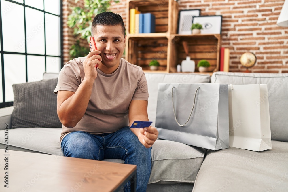 Hispanic young man doing payment with credit card on the phone sticking tongue out happy with funny expression.