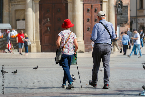 Modern aging couple with a walking stick stroll along a paved road on a sunny summer day. Pigeons and passers-by add diversity and culture to the scene. Nostalgic, authentic ambiance