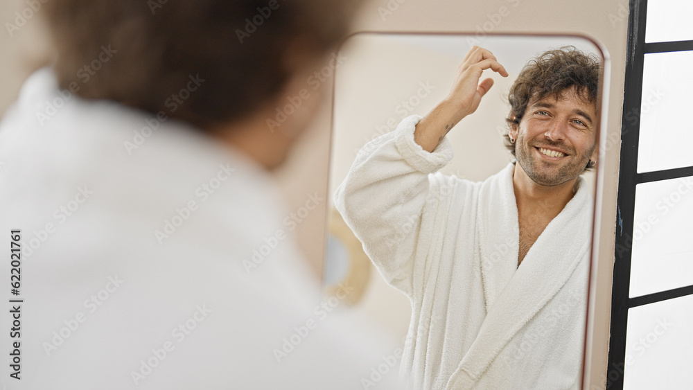 Young hispanic man wearing bathrobe combing hair with hands looking on mirror at home Young hispanic man wearing bathrobe combing hair with hands looking on mirror at home