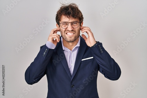 Hispanic business man wearing glasses covering ears with fingers with annoyed expression for the noise of loud music. deaf concept.