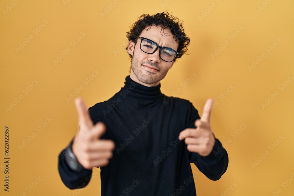 Hispanic man standing over yellow background pointing fingers to camera with happy and funny face. good energy and vibes. Hispanic man standing over yellow background pointing fingers to camera with happy and funny face. good energy and vibes.