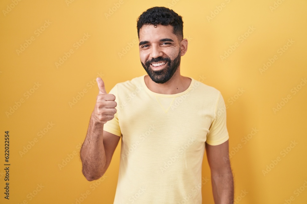Hispanic man with beard standing over yellow background doing happy thumbs up gesture with hand. approving expression looking at the camera showing success.
