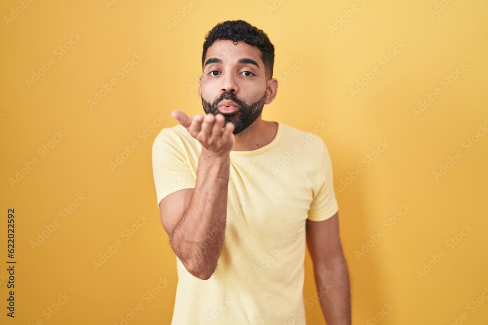Hispanic man with beard standing over yellow background looking at the camera blowing a kiss with hand on air being lovely and sexy. love expression. Hispanic man with beard standing over yellow background looking at the camera blowing a kiss with hand on air being lovely and sexy. love expression.