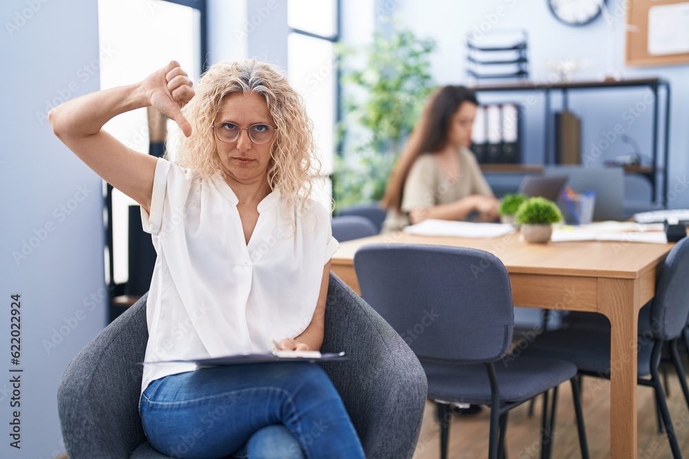 Middle age woman working at the office holding clipboard with angry face, negative sign showing dislike with thumbs down, rejection concept Middle age woman working at the office holding clipboard with angry face, negative sign showing dislike with thumbs down, rejection concept