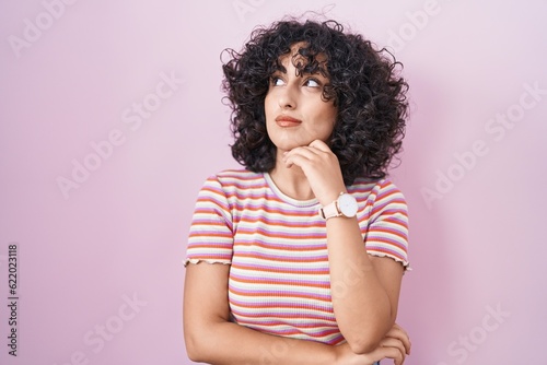 Young middle east woman standing over pink background with hand on chin thinking about question, pensive expression. smiling with thoughtful face. doubt concept.