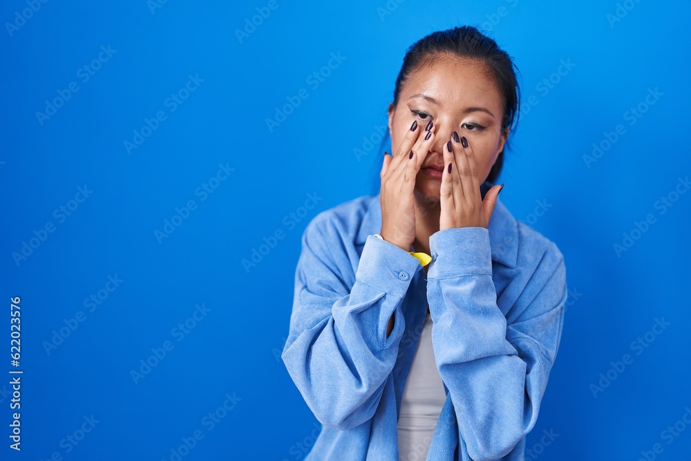 Asian young woman standing over blue background rubbing eyes for fatigue and headache, sleepy and tired expression. vision problem Asian young woman standing over blue background rubbing eyes for fatigue and headache, sleepy and tired expression. vision problem