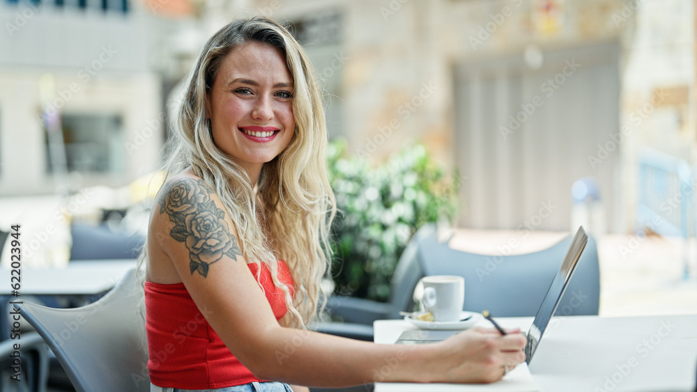 Young blonde woman using laptop taking notes smiling at coffee shop terrace