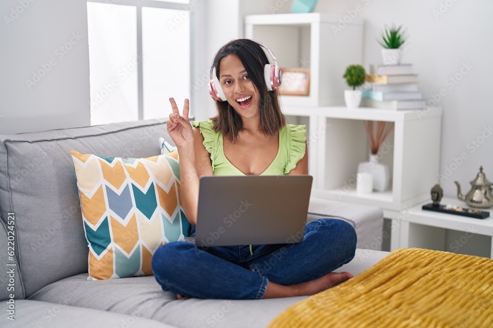 Hispanic young woman using laptop at home smiling with happy face winking at the camera doing victory sign. number two. Hispanic young woman using laptop at home smiling with happy face winking at the camera doing victory sign. number two.