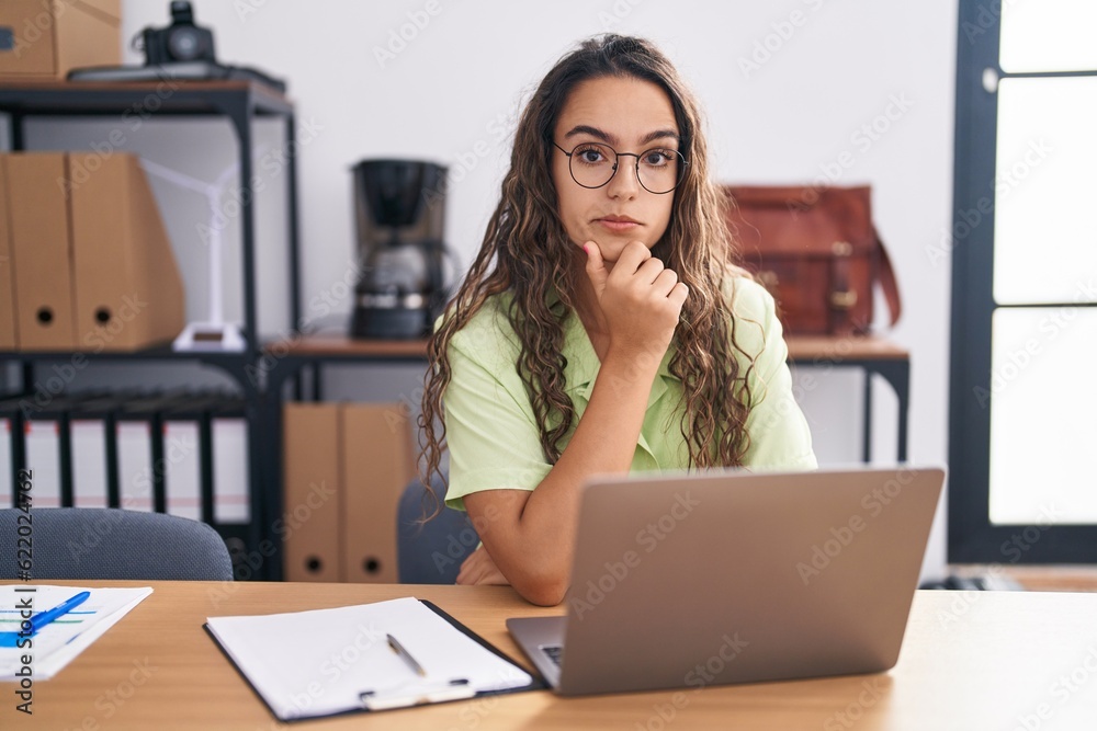Obraz premium Young hispanic woman working at the office wearing glasses looking confident at the camera with smile with crossed arms and hand raised on chin. thinking positive.