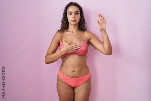Young hispanic woman wearing lingerie over pink background swearing with hand on chest and open palm, making a loyalty promise oath