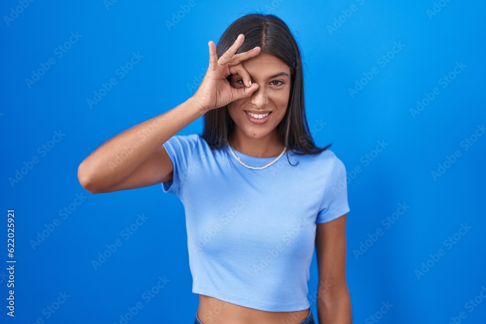 Brunette young woman standing over blue background doing ok gesture with hand smiling, eye looking through fingers with happy face. Brunette young woman standing over blue background doing ok gesture with hand smiling, eye looking through fingers with happy face.