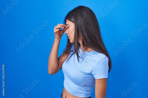 Brunette young woman standing over blue background tired rubbing nose and eyes feeling fatigue and headache. stress and frustration concept.