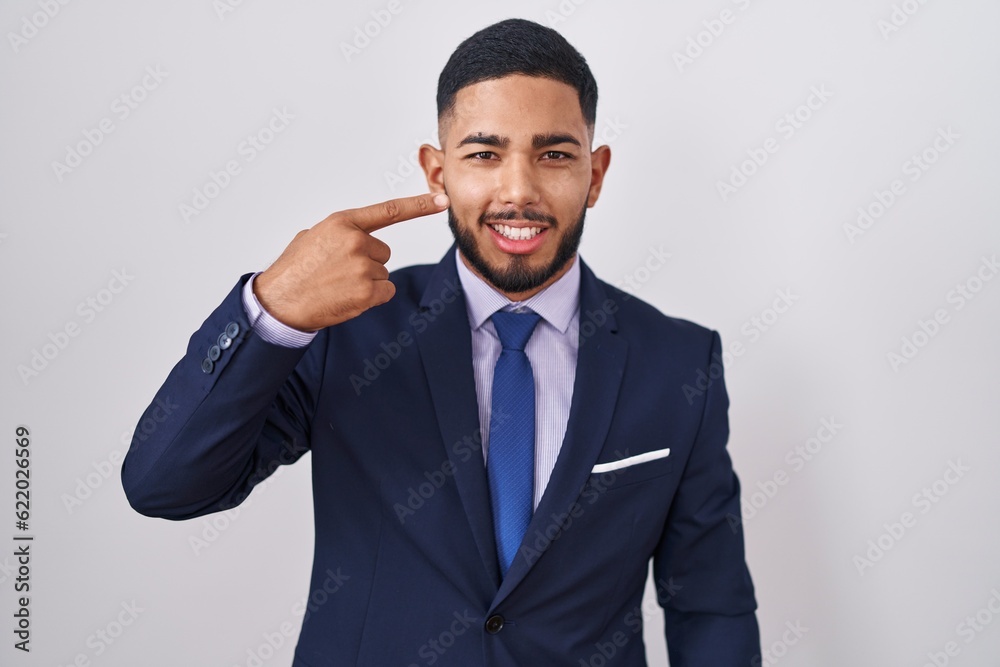 Young hispanic man wearing business suit and tie pointing with hand finger to face and nose, smiling cheerful. beauty concept Young hispanic man wearing business suit and tie pointing with hand finger to face and nose, smiling cheerful. beauty concept