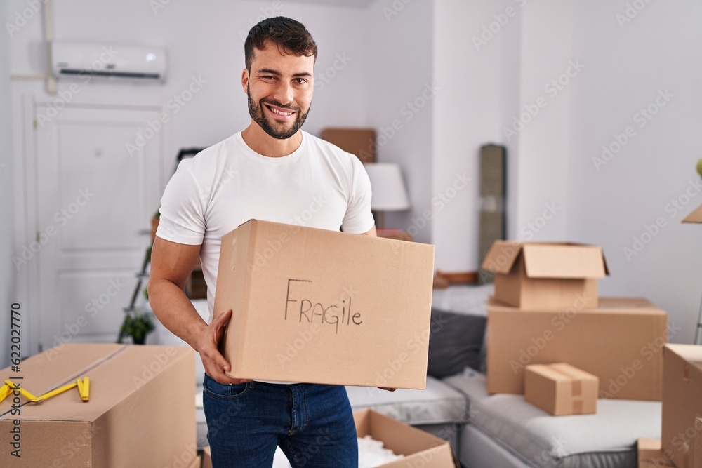 Handsome hispanic man moving to a new home holding fragile box winking looking at the camera with sexy expression, cheerful and happy face. Handsome hispanic man moving to a new home holding fragile box winking looking at the camera with sexy expression, cheerful and happy face.