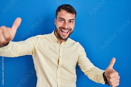 Handsome hispanic man standing over blue background approving doing positive gesture with hand, thumbs up smiling and happy for success. winner gesture.