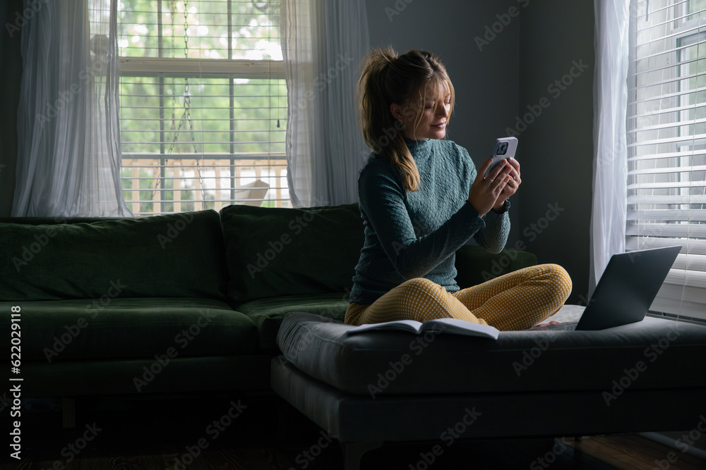 Business Woman Work from Home on Laptop Computer, Remote Employee Worker, Sitting in Living Room of House. Looking at phone screen with grin.