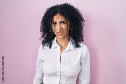 Hispanic woman with curly hair standing over pink background in shock face, looking skeptical and sarcastic, surprised with open mouth