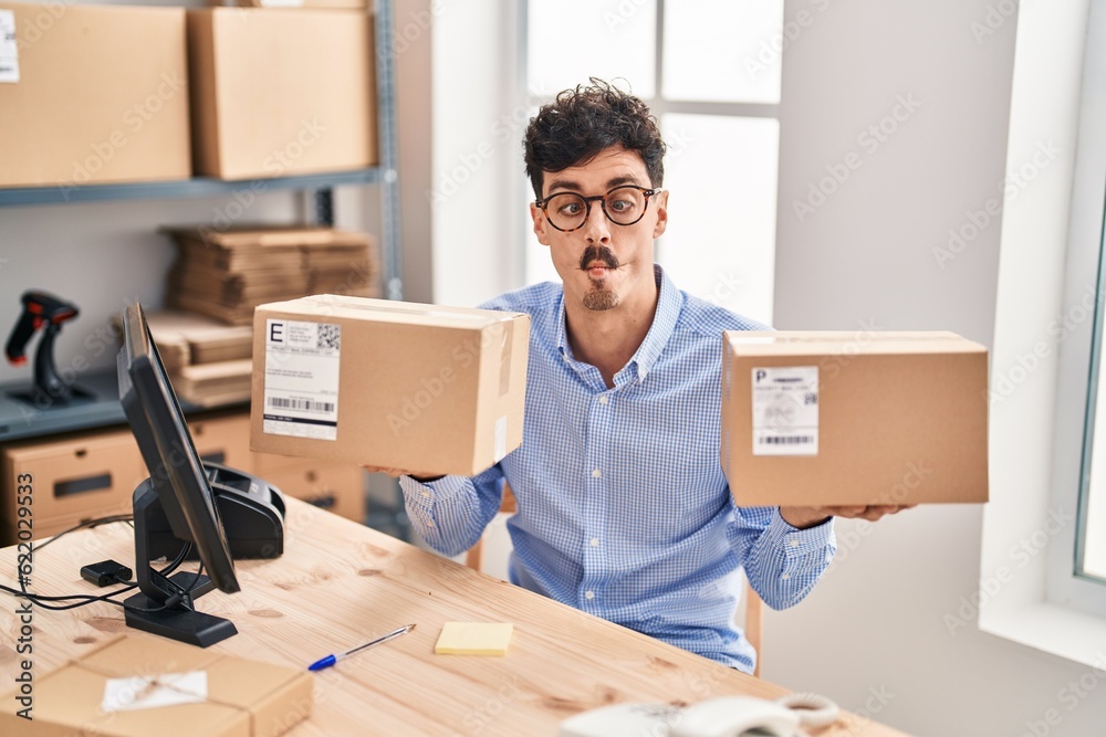 Hispanic man working at small business ecommerce holding packages making fish face with mouth and squinting eyes, crazy and comical. Hispanic man working at small business ecommerce holding packages making fish face with mouth and squinting eyes, crazy and comical.