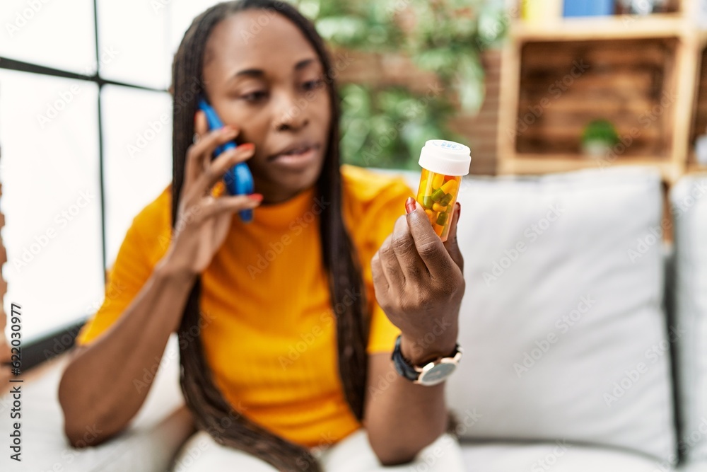 African american woman talking on smartphone with serious expression holding pills at home African american woman talking on smartphone with serious expression holding pills at home