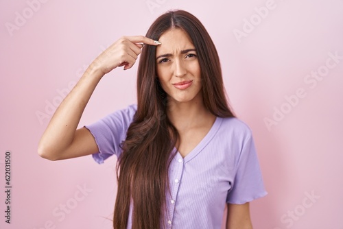 Young hispanic woman with long hair standing over pink background pointing unhappy to pimple on forehead, ugly infection of blackhead. acne and skin problem