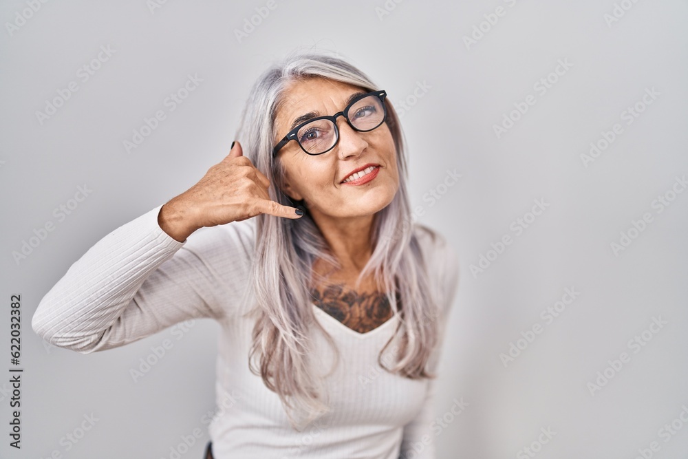Middle age woman with grey hair standing over white background smiling doing phone gesture with hand and fingers like talking on the telephone. communicating concepts. Middle age woman with grey hair standing over white background smiling doing phone gesture with hand and fingers like talking on the telephone. communicating concepts.