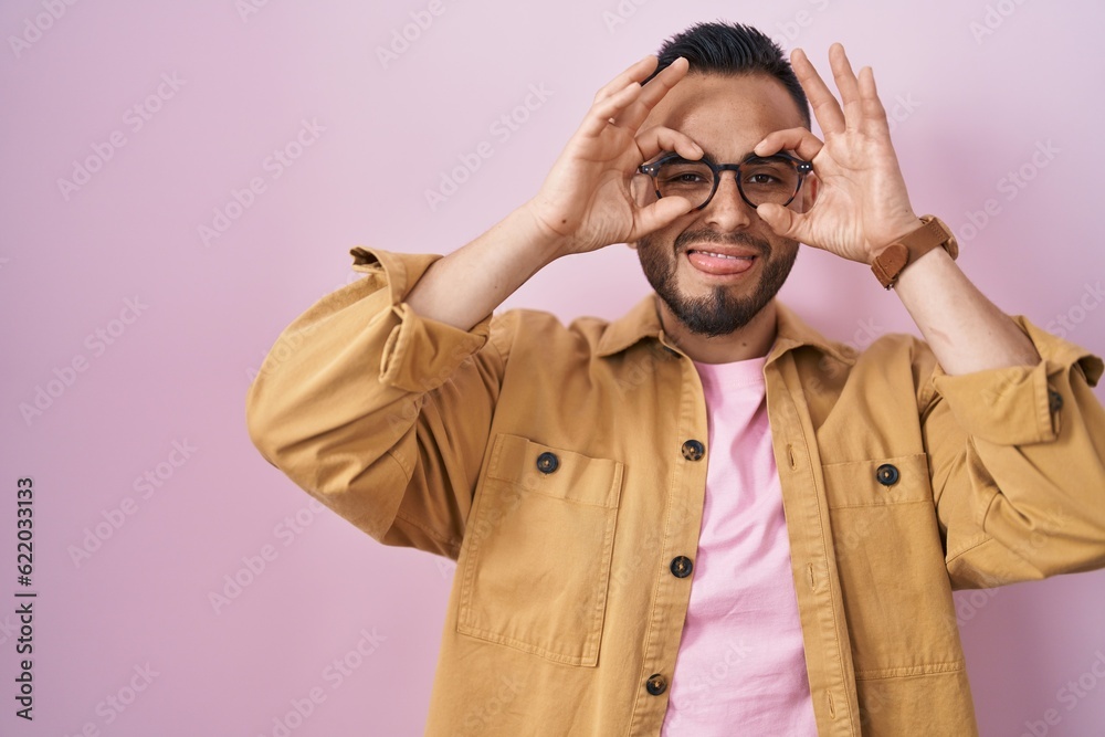 Young hispanic man standing over pink background doing ok gesture like binoculars sticking tongue out, eyes looking through fingers. crazy expression. Young hispanic man standing over pink background doing ok gesture like binoculars sticking tongue out, eyes looking through fingers. crazy expression.