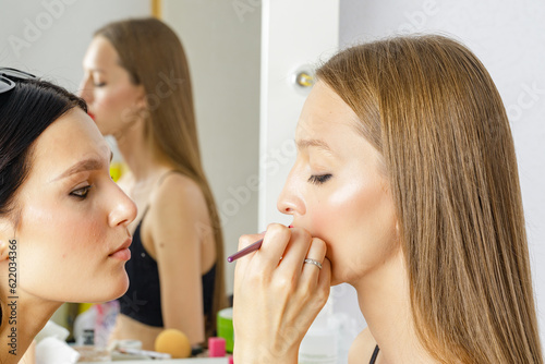 Close up of hand of visagiste applying lipstick on female lips. She is holding a brush. The young beautiful woman with red lip