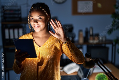 African american woman with braids working at the office at night with tablet waiving saying hello happy and smiling, friendly welcome gesture © Krakenimages.com