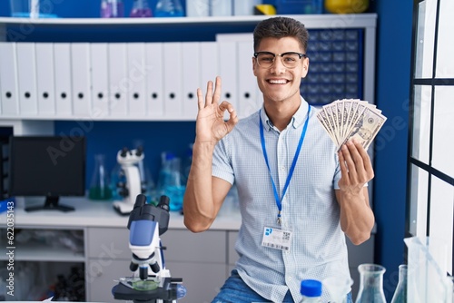 Young hispanic man working at scientist laboratory holding dollars doing ok sign with fingers, smiling friendly gesturing excellent symbol