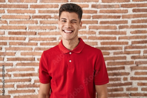 Young hispanic man standing over bricks wall winking looking at the camera with sexy expression, cheerful and happy face.