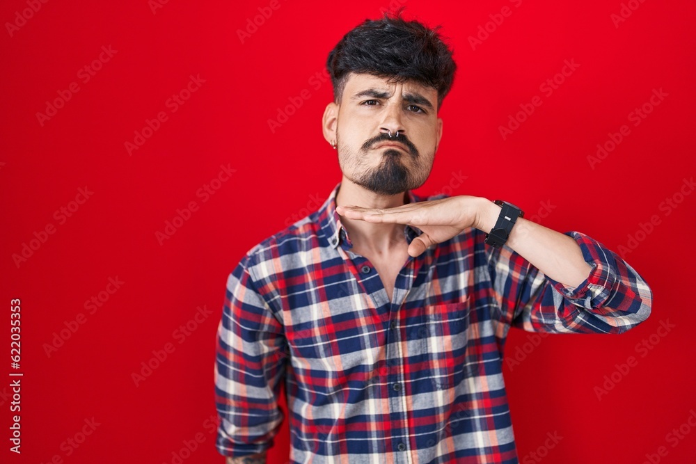 Young hispanic man with beard standing over red background cutting throat with hand as knife, threaten aggression with furious violence