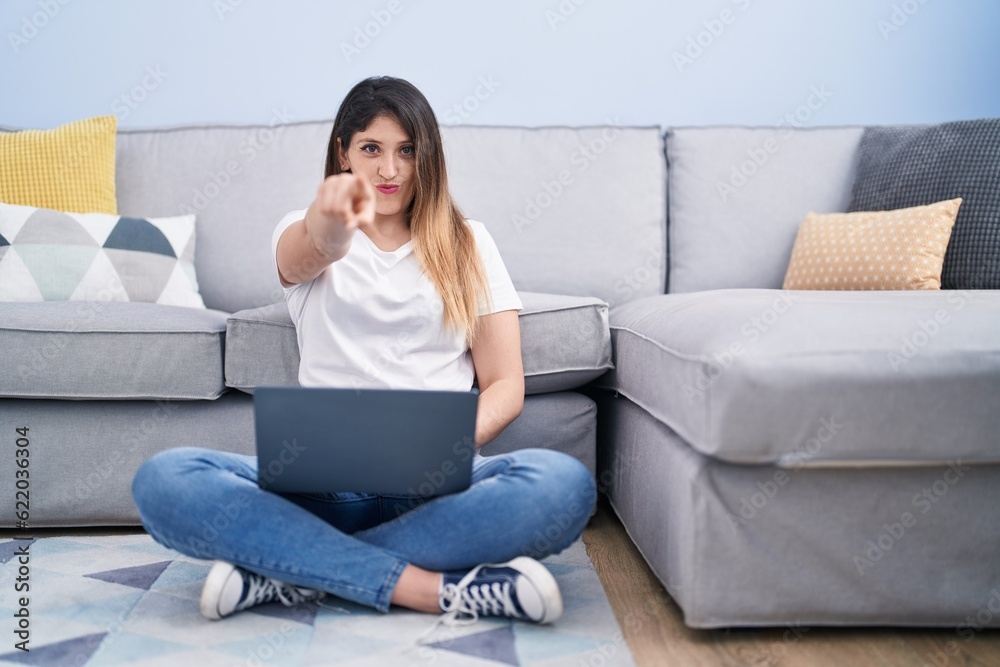 Young brunette woman sitting on the floor at home using laptop pointing with finger to the camera and to you, confident gesture looking serious Young brunette woman sitting on the floor at home using laptop pointing with finger to the camera and to you, confident gesture looking serious