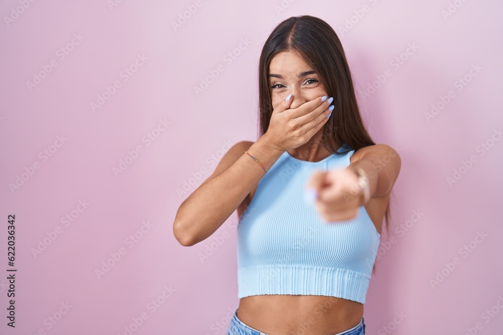Young brunette woman standing over pink background laughing at you, pointing finger to the camera with hand over mouth, shame expression