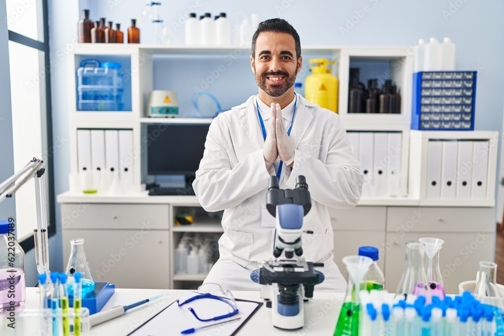 Young hispanic man with beard working at scientist laboratory praying with hands together asking for forgiveness smiling confident. Young hispanic man with beard working at scientist laboratory praying with hands together asking for forgiveness smiling confident.
