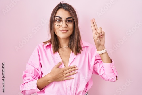 Young hispanic woman wearing glasses standing over pink background smiling swearing with hand on chest and fingers up  making a loyalty promise oath