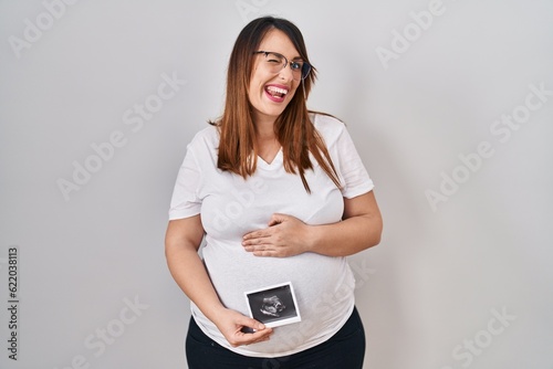 Pregnant woman holding baby ecography winking looking at the camera with sexy expression, cheerful and happy face.