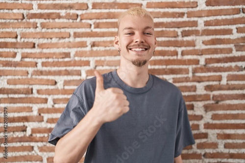 Young caucasian man standing over bricks wall doing happy thumbs up gesture with hand. approving expression looking at the camera showing success.