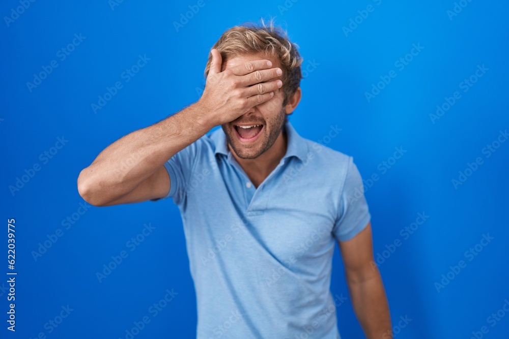 Caucasian man standing over blue background smiling and laughing with hand on face covering eyes for surprise. blind concept. Caucasian man standing over blue background smiling and laughing with hand on face covering eyes for surprise. blind concept.