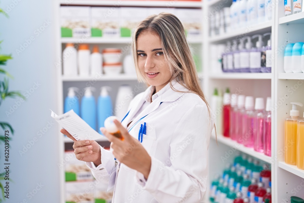 Young blonde woman pharmacist holding pills bottle reading prescription at pharmacy