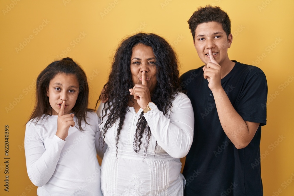 Family of mother, daughter and son standing over yellow background asking to be quiet with finger on lips. silence and secret concept. Family of mother, daughter and son standing over yellow background asking to be quiet with finger on lips. silence and secret concept.