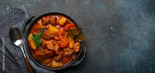 Beef meat stew with potatoes, carrot and delicious gravy in black casserole pot with bay leaves and fresh green herbs with spoon on dark rustic concrete background from above, space for text.